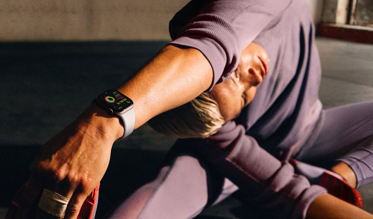 Une femme assise sur le sol étire son bras vers sa jambe; elle porte une Apple Watch Series 10 ; l’écran affiche Exercice, Récupération, Activité, Anneaux Activité