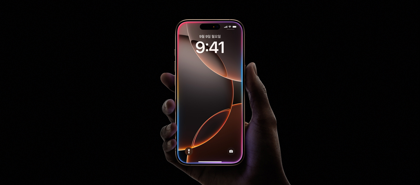 데저트 티타늄 색상의 iPhone 16 Pro를 한 손으로 잡고 있는 모습. 전면 화면 디스플레이, 상단 중앙의 Dynamic Island, 둥근 형태의 모서리가 보입니다.