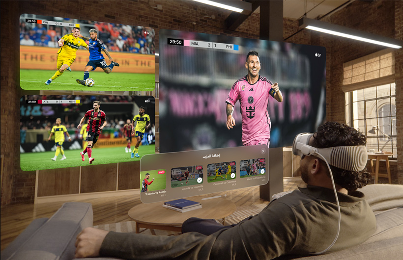 A man wearing Apple Vision Pro is sitting on a couch in a living room. Displayed in front of him are a large video screen alongside two smaller screens, each showing a different sporting event. Below the large screen is selection window that reads "Add More" along the top