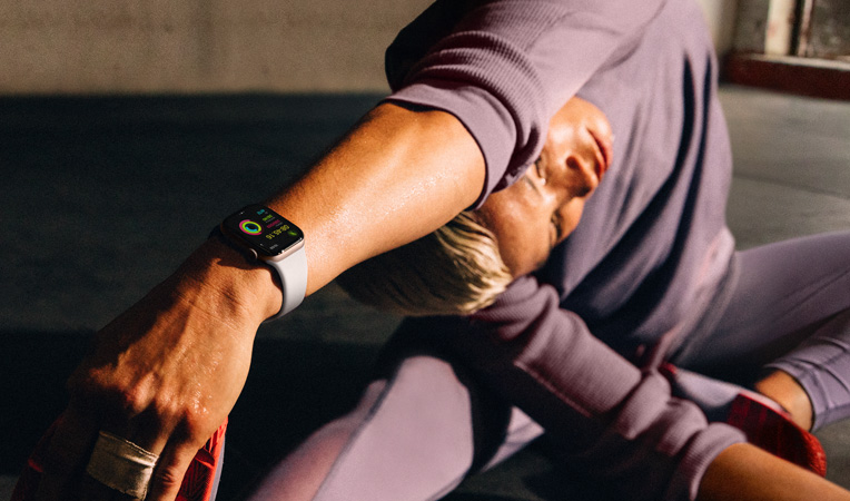 ผู้หญิงนั่งบนพื้นอยู่ในท่าเหยียดแขนพร้อมโน้มตัวไปแตะขากำลังสวม Apple Watch Series 10, หน้าจอแสดงแอปออกกำลังกาย, คูลดาวน์, กิจกรรม, วงแหวนกิจกรรม