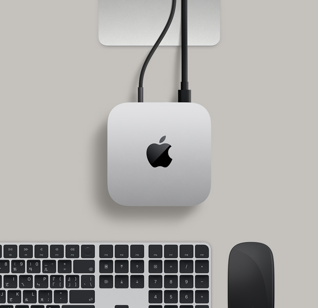 銀色 Mac mini 的俯視圖,兩條插入背面連接埠的連接線連著一部顯示器,旁邊放著黑色巧控滑鼠和黑色巧控鍵盤