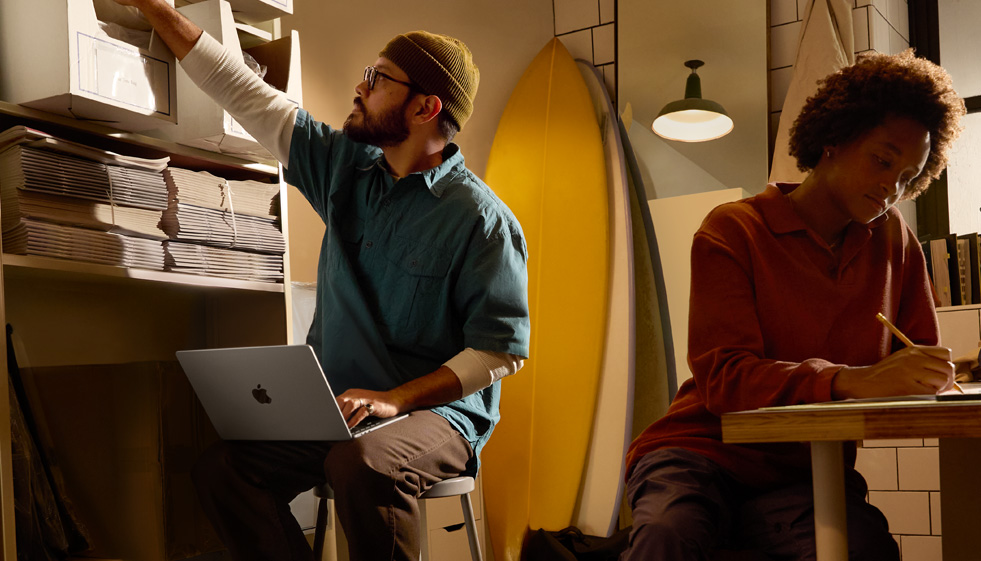 شخص في بيئة عمل يجلس على مقعد من دون ظهر ويستخدم جهاز MacBook Air غير موصول بالطاقة وموضوع على ركبتيه بينما يمد يده الأخرى للوصول إلى علبة على رف عال